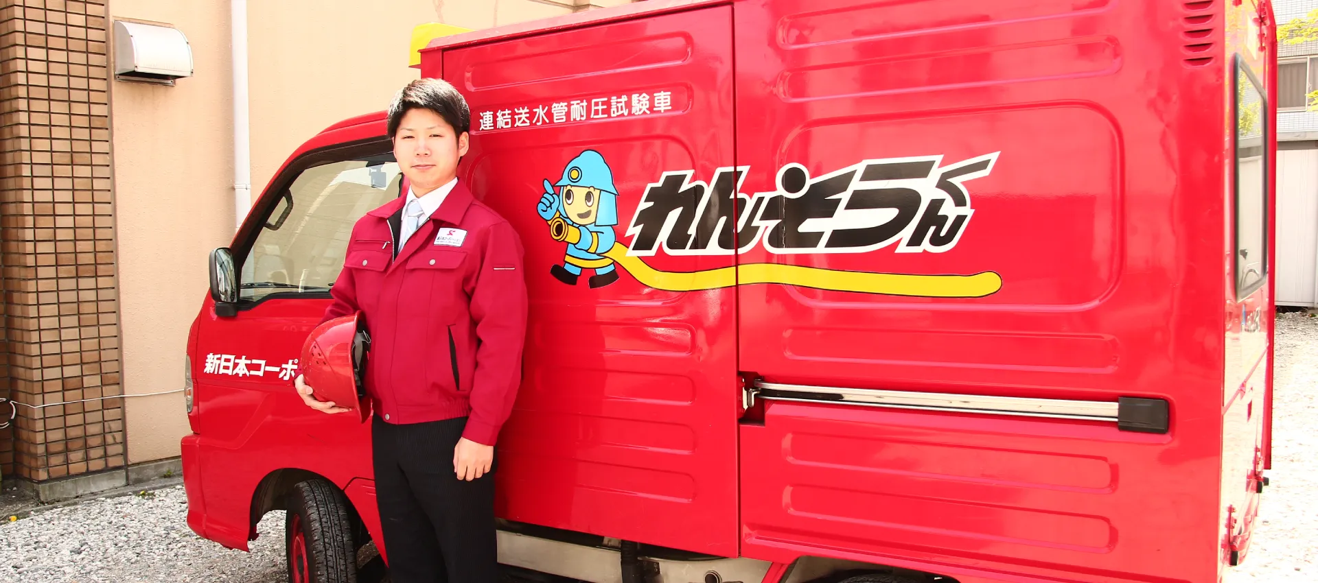 連結送水管耐圧試験専用車を背景に、赤いヘルメットを抱えている男性の写真